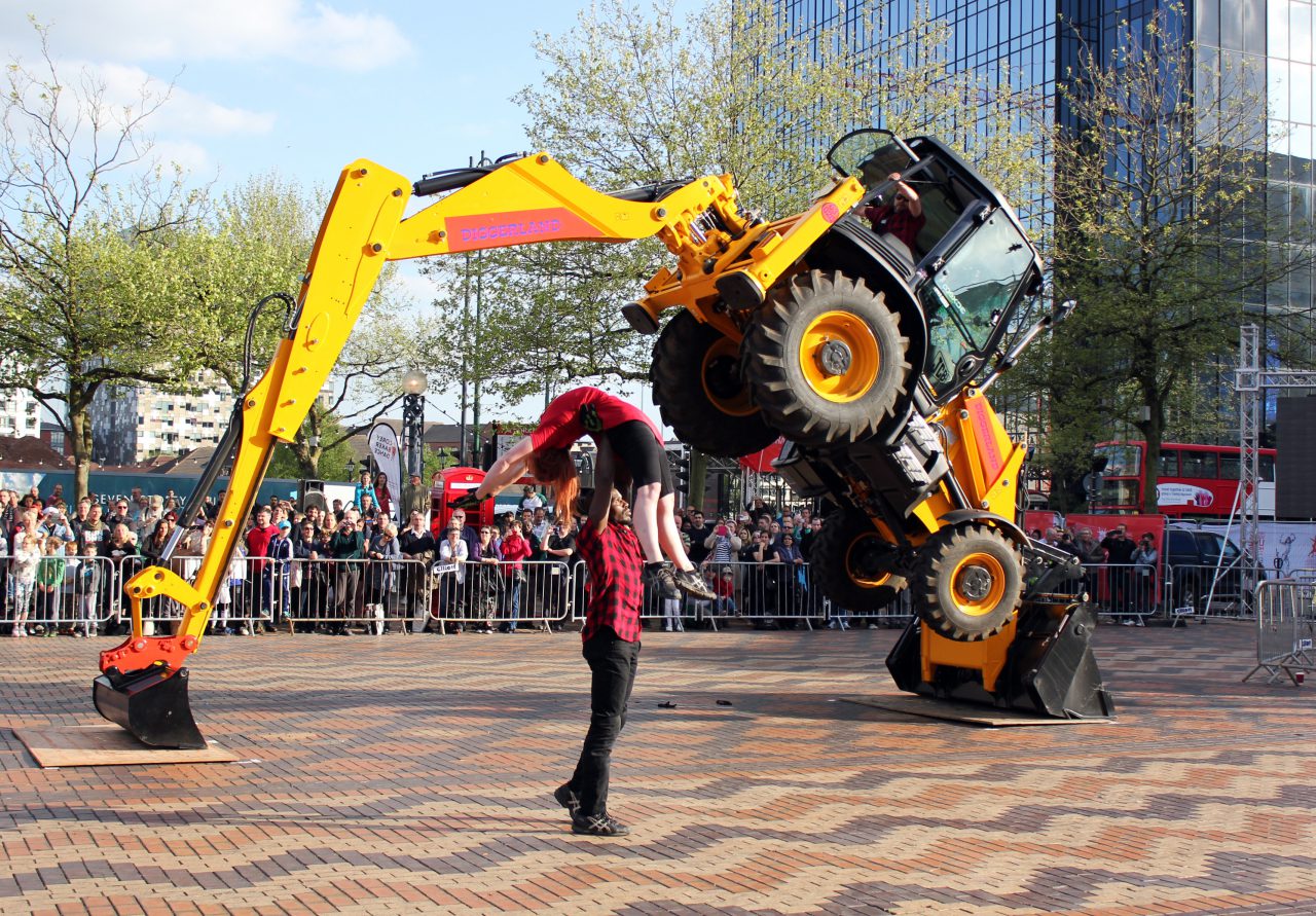 Man lifts woman over his head with one arm. JCB digger in background balanced on its rear bucket
