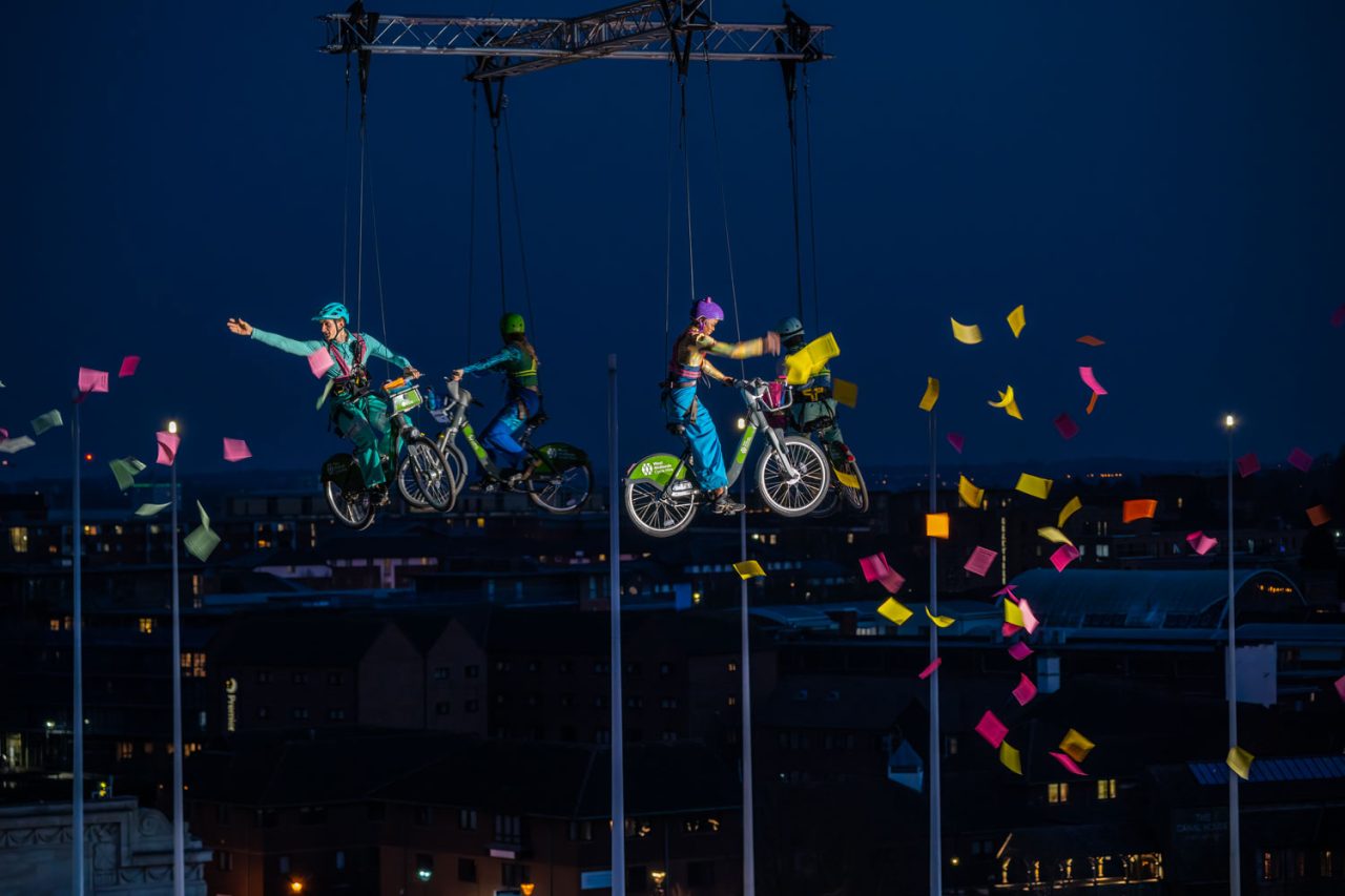 4 people riding bicycles that appear to be flying across the night sky, throwing colourful paper that flutters to the floor