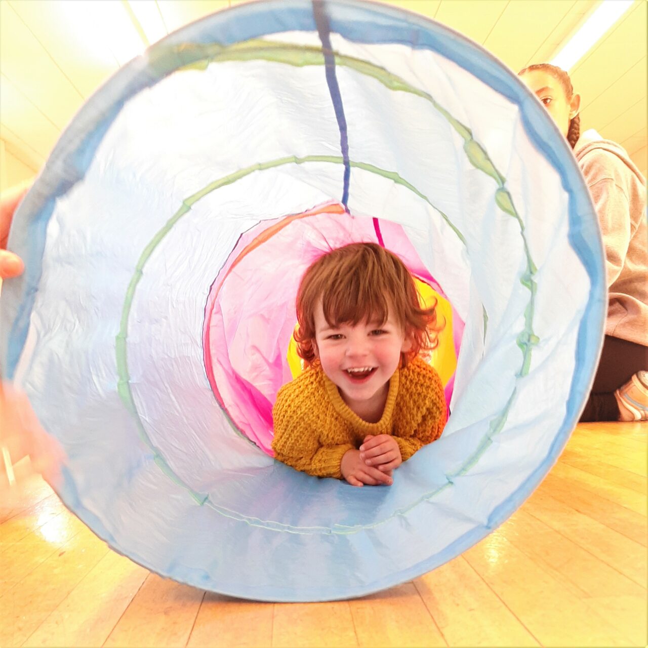 Young boy in yellow jumper smiles as he crawls through a colourful play tunnel