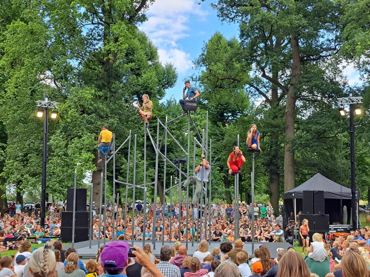 A group of people sit on top of tall poles while a large crowd watches below