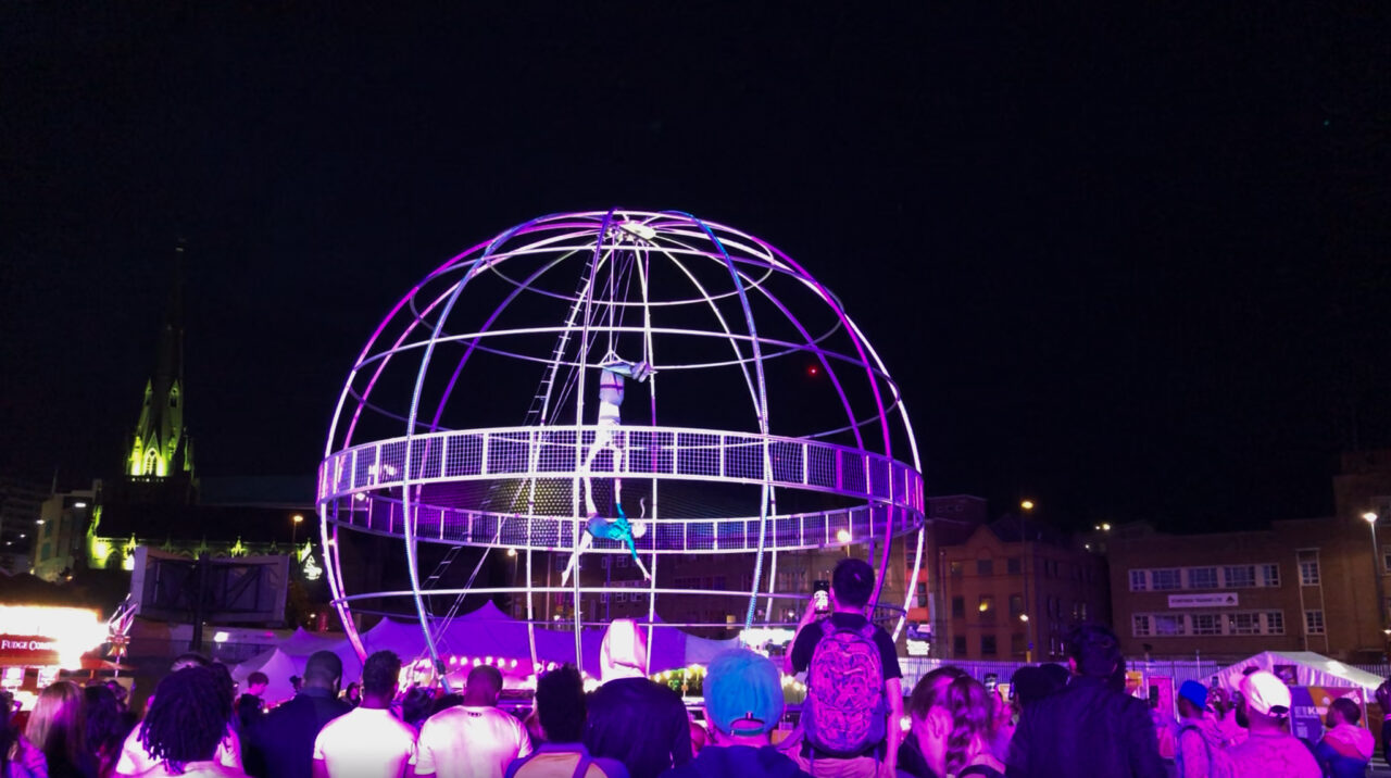 Two dancers are inside a large metal globe and dangle from the top of the globe